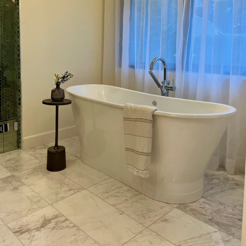 A freestanding white bathtub with a striped towel draped over the side, next to a small side table with a vase of flowers. A shower area with green mosaic tiles is visible on the left.